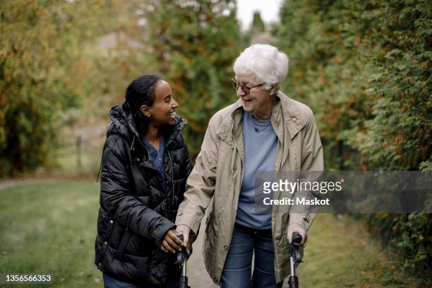 happy female caregiver talking with senior woman while walking in park - asistente sanitario fotografías e imágenes de stock
