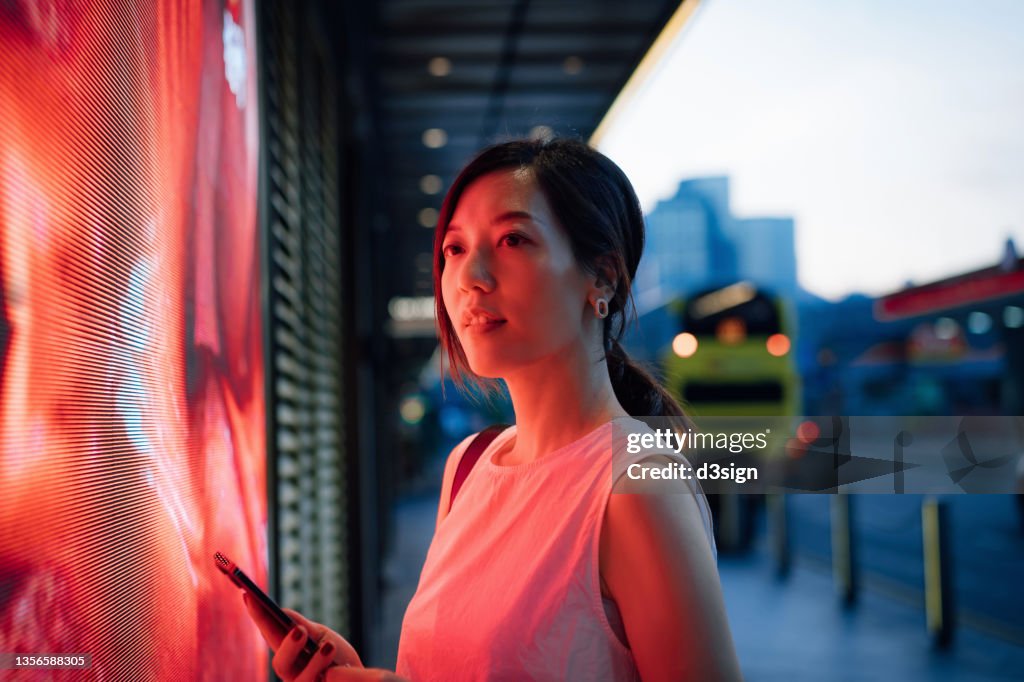 Confident young Asian businesswoman standing against illuminated LED digital display in the city, lit by red neon coloured lights, using smartphone in downtown city street in the evening. Lifestyle and technology