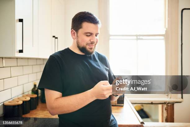 happy man standing in the kitchen and texting on his cell phone - smirking stock pictures, royalty-free photos & images