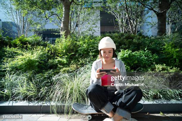 asian female enjoying some entertainment on her smartphone with a skateboard. - asian watching movie stock pictures, royalty-free photos & images