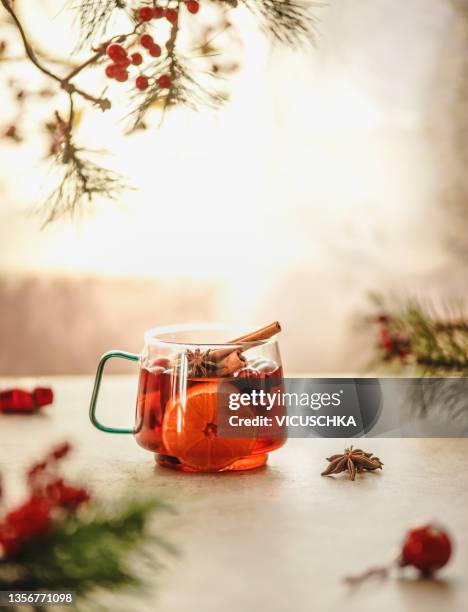 winter punch with orange, chinnamon and star anise in glass mug on kitchen table with fir green at window background with natural light - bowle stock-fotos und bilder