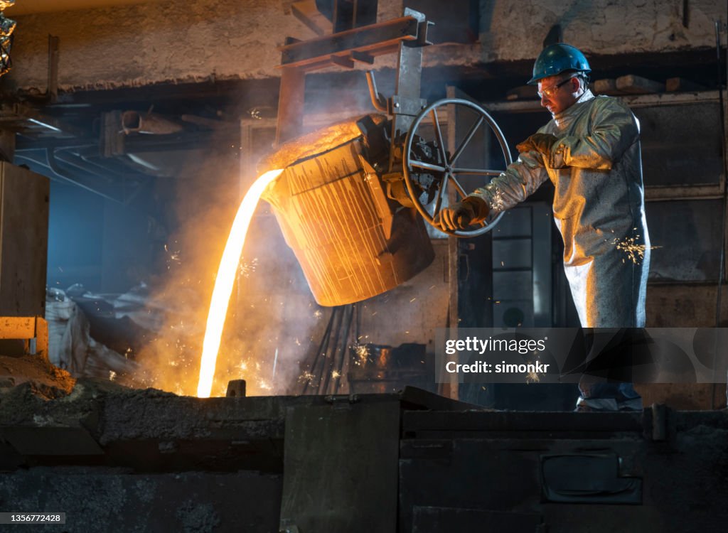 Foundry worker in protective coat pouring molten metal from the ladle