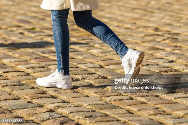 Passerby wears a white long coat, navy blue denim skinny jeans pants, white leather sneakers, on November 29, 2021 in Paris, France.