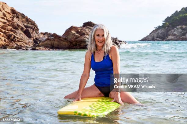 mature woman surfing on the mediterranian sea with a yellow surfboard. - women in bathing suits stock pictures, royalty-free photos & images