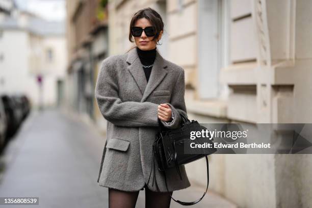 Katie Giorgadze @katie.one wears black sunglasses, a black ribbed wool turtleneck pullover, a silver chain necklace, a gray wool oversized coat, a...