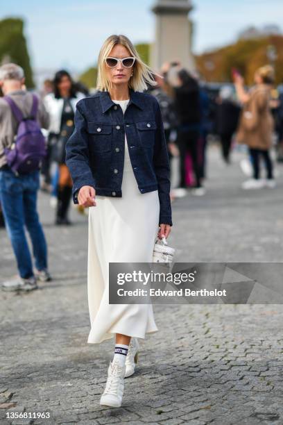 Guest wears white sunglasses, gold and pearls earrings, a navy blue denim jacket, white midi dress, a white Vanity case handbag from Dior, white...