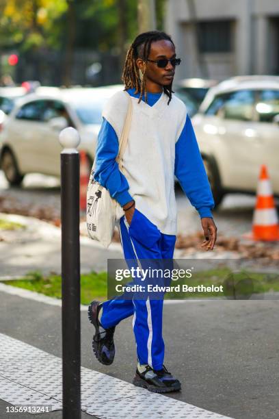 Guest wears sunglasses, a royal blue pullover, a white braided wool / sleeveless oversized pullover, a white with black inscriptions print pattern...