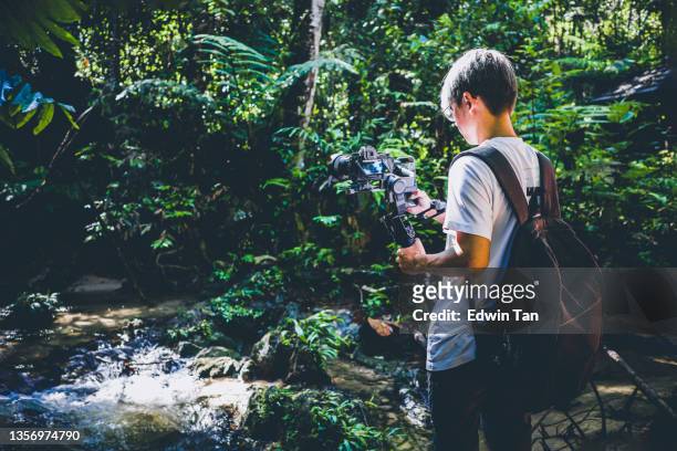rückansicht asiatischer chinesischer teenager-kameramann, der am wochenendmorgen mit spiegelloser kamera und gimbal ein video im regenwaldufer aufnimmt - boy taking picture in forest stock-fotos und bilder