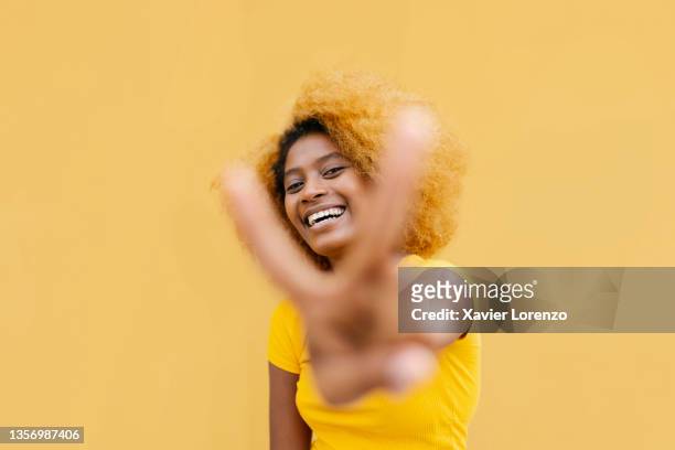 happy young latina woman showing peace gesture in front of yellow wall - friedenszeichen handzeichen stock-fotos und bilder