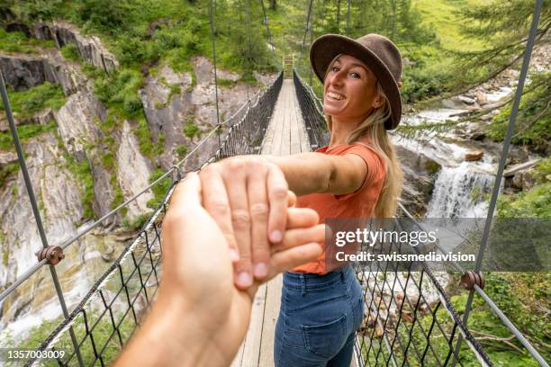 follow me konzept: junge frau, die die hand des begleiters hält und ihn über eine hängebrücke im wald führt. wanderpaar händchen haltend - woman hand crossed stock-fotos und bilder