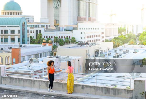 two cool young women in vibrant clothing hang out together on an urban summer rooftop overlooking the city of west palm beach florida - west palm beach stock pictures, royalty-free photos & images