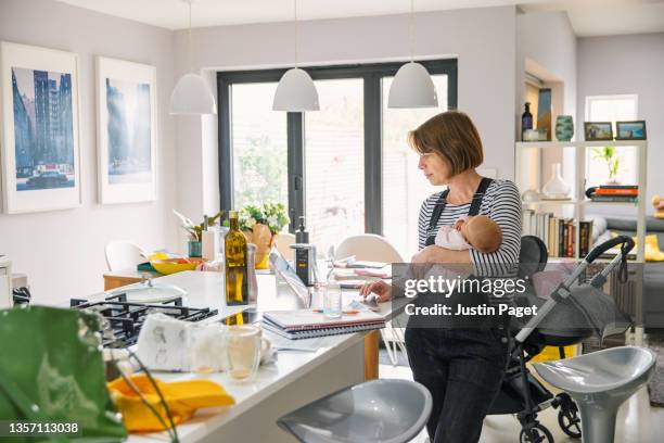 mature woman working from home whilst holding her newborn baby daughter - homemaker stock pictures, royalty-free photos & images