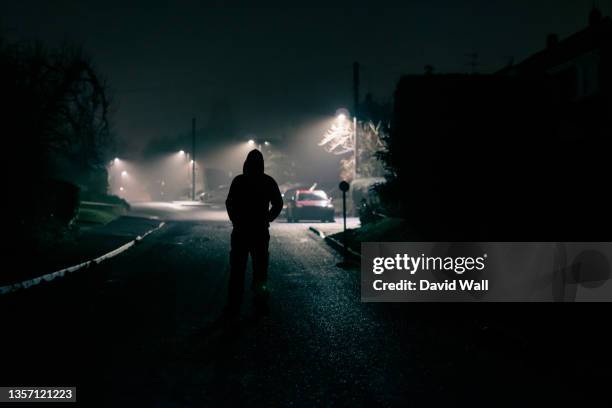 a moody concept of a scary hooded figure. standing in the middle of the road in a quiet suburban street. on a foggy winters night - mystery crime stock pictures, royalty-free photos & images