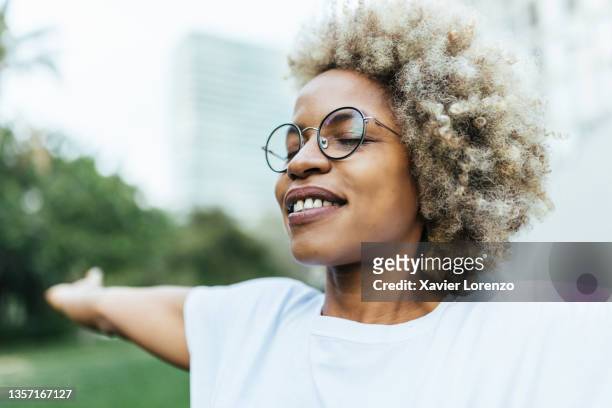 smiling afro woman with closed eyes and open arms enjoying outdoors - brille frühling stock-fotos und bilder