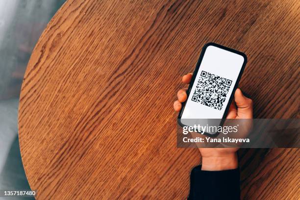 man hand holding a mobile phone with digital vaccination qr code or vaccine passport over a wooden cafe table - qr stockfoto's en -beelden