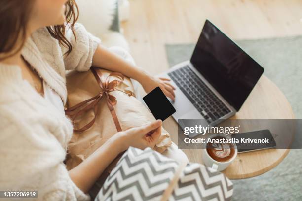 woman sitting on sofa with gifts and using computer and credit card for online shopping. - phone credit card stock-fotos und bilder