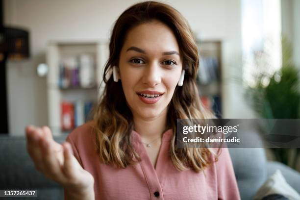 giovane donna sorridente con cuffie bluetooth che ha una videochiamata a casa - looking at phones foto e immagini stock