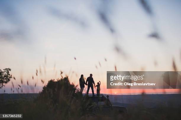 couple with child standing on car and seeing off the sunset. travel and family adventure concept - great american group stock pictures, royalty-free photos & images