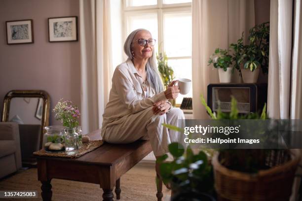 fashionable senior woman sitting on table and drinking coffee at home. - camel active fotografías e imágenes de stock