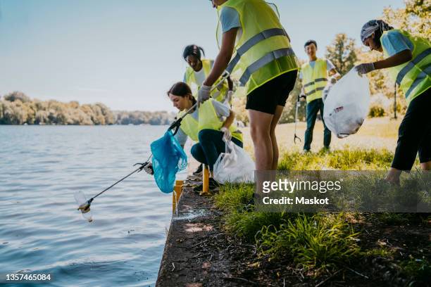 young female and male environmentalist cleaning lake while picking plastic from claw - volunteer beach photos et images de collection