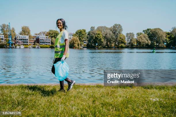 portrait of young woman walking while holding plastic bag by lake - volunteer beach photos et images de collection
