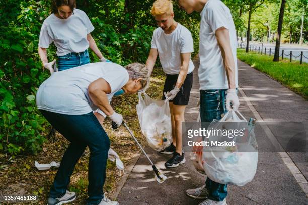 senior female activist picking up face mask while cleaning park with young volunteers - generation z covid stock pictures, royalty-free photos & images