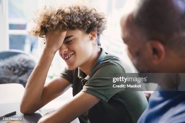 smiling teenage boy with hand in hair sitting by father at home - boy talking stock pictures, royalty-free photos & images