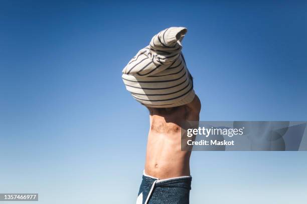 man taking off t-shirt against clear sky on sunny day - spogliarsi foto e immagini stock
