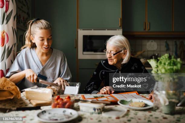 female caregiver cutting bread loaf at dining table while sitting by senior woman in kitchen - old man young woman stock-fotos und bilder