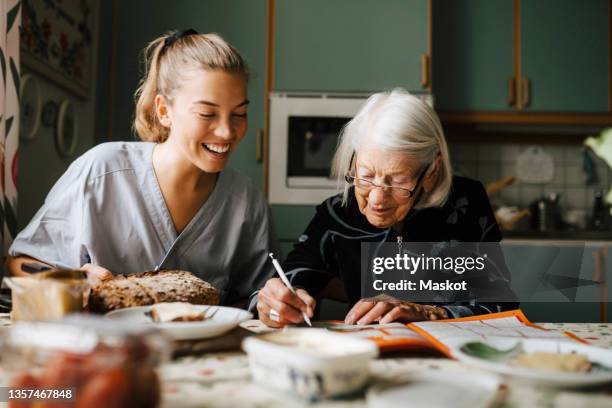 senior woman solving crossword puzzle in book sitting by smiling nurse in kitchen at home - carer stock-fotos und bilder