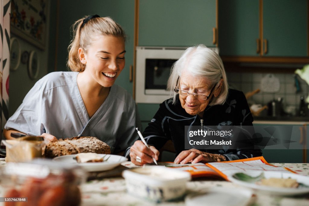 Senior woman solving crossword puzzle in book sitting by smiling nurse in kitchen at home