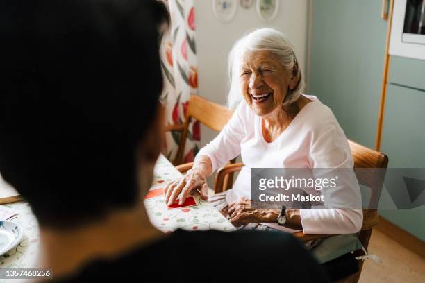 cheerful senior woman talking with male caregiver while playing cards in kitchen - asistencia de la comunidad fotografías e imágenes de stock