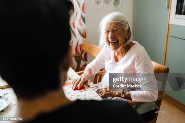 cheerful senior woman talking with male caregiver while playing cards in kitchen - mais de 90 anos - fotografias e filmes do acervo
