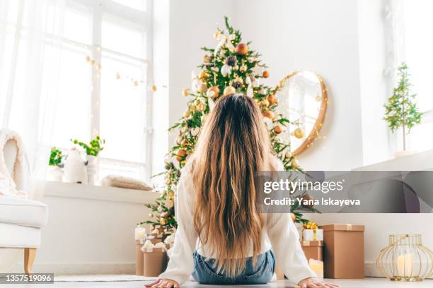 beautiful woman with long wavy hair is sitting on floor enjoying the decorated christmas tree, rear view - kerstboom versieren stockfoto's en -beelden