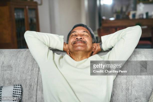 shot of a happy senior man relaxing on the sofa at home - hands behind head stockfoto's en -beelden