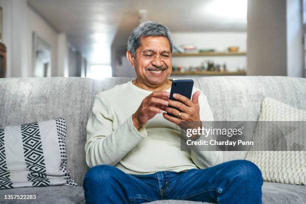 foto de un hombre mayor usando un teléfono inteligente en el sofá de su casa - hombres-mayores fotografías e imágenes de stock