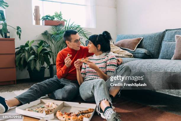 a couple enjoying eating pizza while sitting on the floor of their cozy living room - sharing pizza stock pictures, royalty-free photos & images