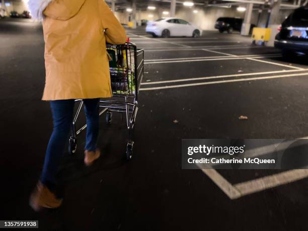 young female wearing bright mustard yellow winter coat walks briskly pushing grocery shopping cart in parking garage - parking space stock-fotos und bilder