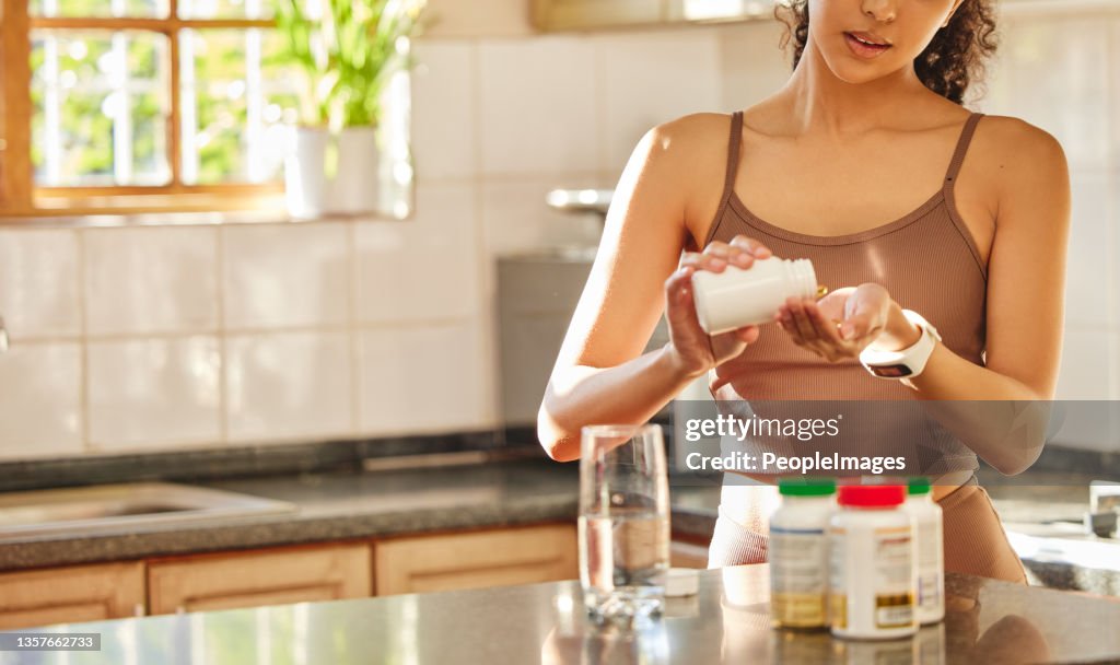 Cropped shot of an unrecognisable woman standing alone in her kitchen and taking her vitamins