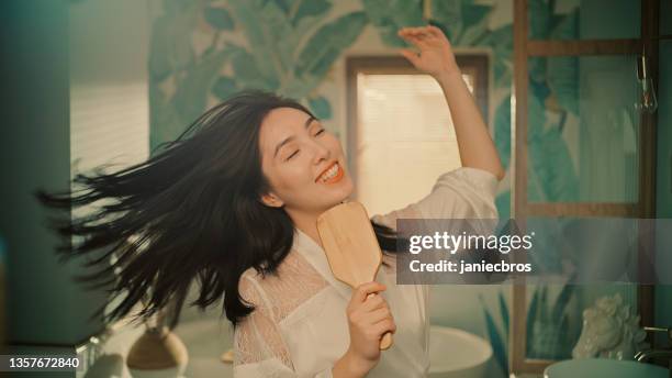 feeling fabulous. asian ethicity woman enjoying morning bathroom routine and having fun in front of mirror. singing to a hairbrush - woman brushing hair stockfoto's en -beelden