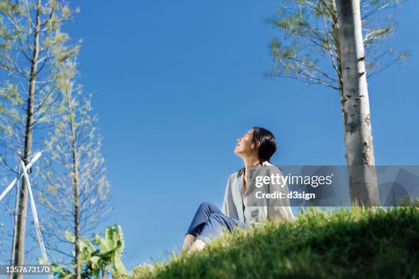 carefree young asian woman with eyes closed relaxing in park, sitting on meadow against blue sky, enjoying the warmth of sunlight on a relaxing afternoon in nature on a beautiful sunny day - woman fresh air photos et images de collection