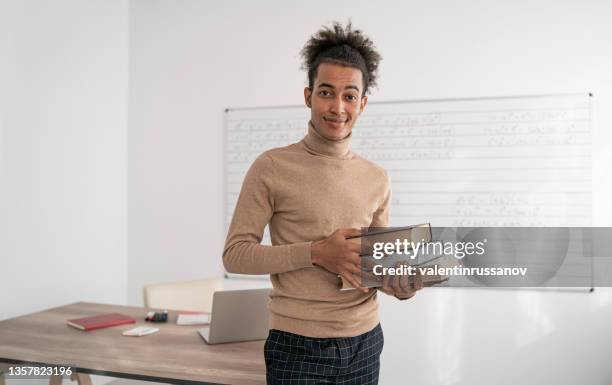 an african male teacher standing in an empty classroom and holding books - closing book stock pictures, royalty-free photos & images