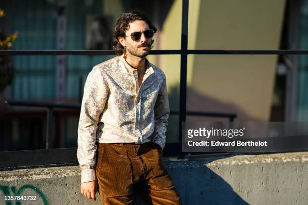 Guest wears sunglasses from Ray Ban, a gold with brown pompom pendant earring, a white latte with gold print patter shirt, brown ribbed velvet pants,...