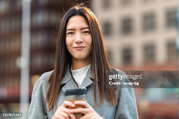 porträt einer schönen gemischtrassigen geschäftsfrau, die in der stadt eine wiederverwendbare kaffeetasse hält und für die kamera lächelt - mixed race woman drinking tea stock-fotos und bilder