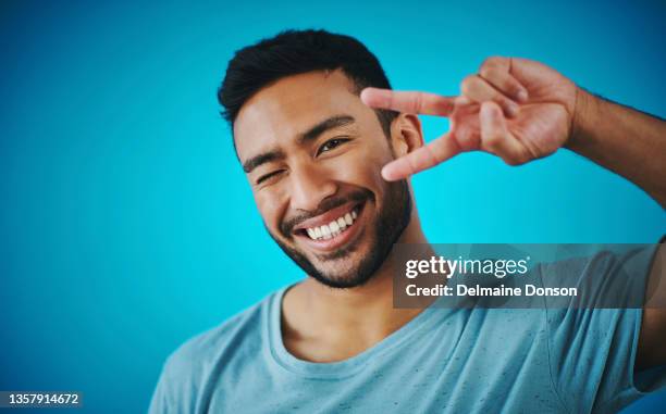 shot of a handsome young man showing the peace sign against a blue background - peace sign guy stock pictures, royalty-free photos & images