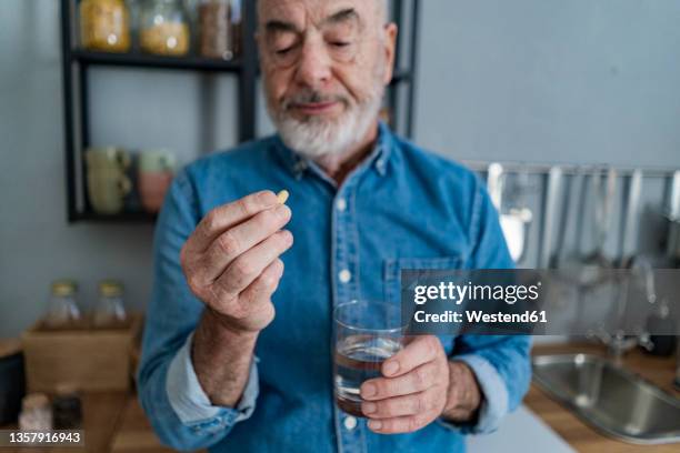 retired man holding glass of water looking at pill in kitchen - taking medication stockfoto's en -beelden