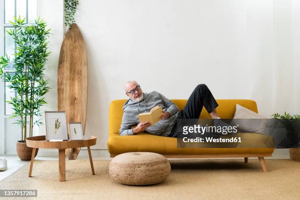 senior man reading book and resting on sofa at home - lying on side stockfoto's en -beelden