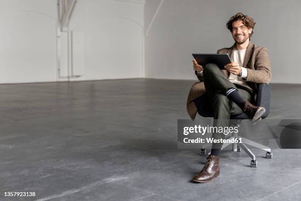 smiling businessman with digital tablet sitting on chair in empty industrial hall - bürostuhl stock-fotos und bilder