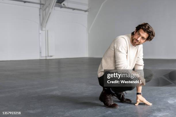 smiling businessman crouching in industrial hall - hurken stockfoto's en -beelden