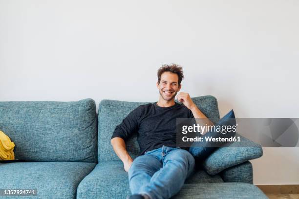 happy young man sitting on sofa in living room at home - wohnzimmer frontal stock-fotos und bilder
